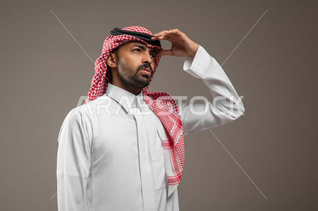 Supporting people and providing assistance, facial and hand gestures indicating boredom and boredom, expressions of respect and appreciation, a close-up portrait of a Saudi Gulf Arab citizen wearing the Saudi thobe and shemagh, placing his hand on his head, looking at something, gray background, sensitive content, sensitive use.