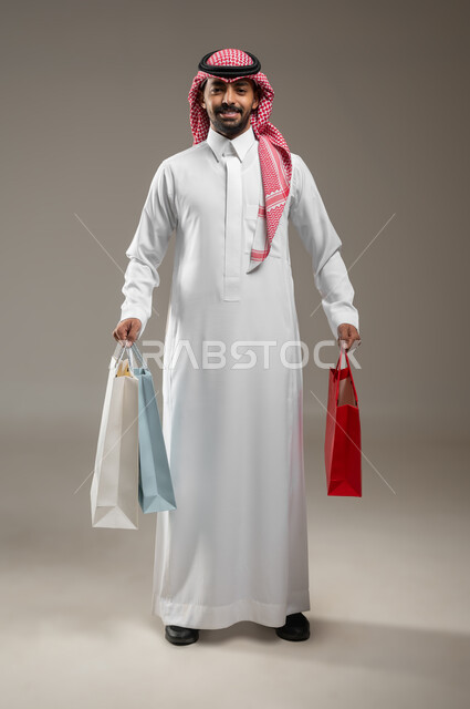 تعابير السرور والسعادة، حسومات وعروض الجمعة البيضاء في السعودية، رجل عربي خليجي سعودي مبتسم يرتدي الشماغ والثوب التقليدي يحمل بيده أكياس ورقية ملونة، متعة التسوق واقتناء المستلزمات والهدايا، بورتريه بالطول الكامل، خلفيه رماديه، محتوى حساس