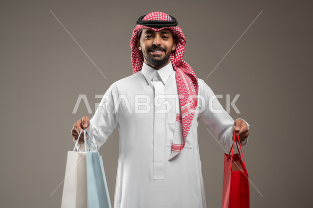 تعابير السرور والسعادة، حسومات وعروض الجمعة البيضاء في السعودية، رجل عربي خليجي سعودي مبتسم يرتدي الشماغ والثوب التقليدي يحمل بيده أكياس ورقية ملونة، متعة التسوق واقتناء المستلزمات والهدايا، بورتريه مقرب، خلفيه رماديه، محتوى حساس