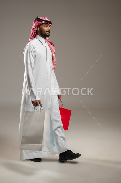 شراء الأغراض ومتعة التسوق في المولات والمحلات التجارية، تعابير وحركات الحماس للتسوق واقتناء المستلزمات والهدايا، بورتريه بالطول الكامل لرجل عربي خليجي سعودي مبستم يرتدي الغترة والثوب السعودي يسير و يحمل بيده أكياس ورقية ملونة، خلفية رماديه، محتوى حساس