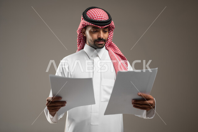 بورتريه مقرب لرجل عربي خليجي سعودي يرتدي الزي السعودي يحمل بيديه اوراق بيضاء، متابعة المراسلات والسجلات ،انجاز المهام المطلوبة، ينظر الى الاوراق بتمعن، التركيز والتدقيق، خلفيه رماديه، استخدام حساس، محتوى حساس