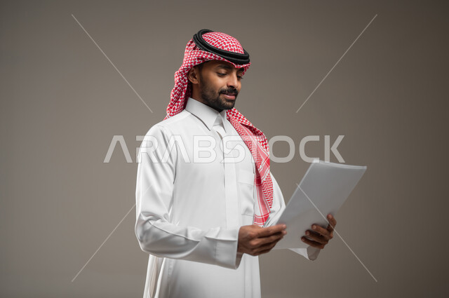 انجاز المهام المطلوبة، بورتريه جانبي لرجل عربي خليجي سعودي يرتدي الزي السعودي يحمل بيده اوراق بيضاء، متابعة المراسلات والسجلات، ينظر الى الاوراق بتمعن، التركيز والتدقيق، خلفيه رماديه، استخدام حساس، محتوى حساس
