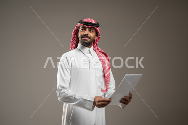 النظر إلى الاعلى والبحث عن شئ ما, بورتريه مقرب لشاب عربي خليجي سعودي مبتسم يرتدي الثوب والشماغ السعودي يمسك بيده ورقه بيضاء يرفع راسه للاعلى للنظر الى شئ ما, ايماءات النظر الى مكان ما، خلفيه رماديه، محتوى حساس، استخدام حساس