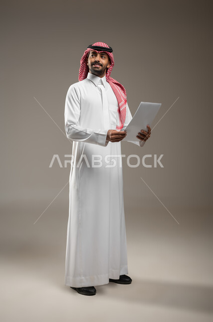 النظر إلى الاعلى والبحث عن شئ ما, بورتريه بالطول الكامل لشاب عربي خليجي سعودي مبتسم يرتدي الثوب والشماغ السعودي يمسك بيده ورقه بيضاء يرفع راسه للاعلى للنظر الى شئ ما, ايماءات النظر الى مكان ما، خلفيه رماديه، محتوى حساس، استخدام حساس