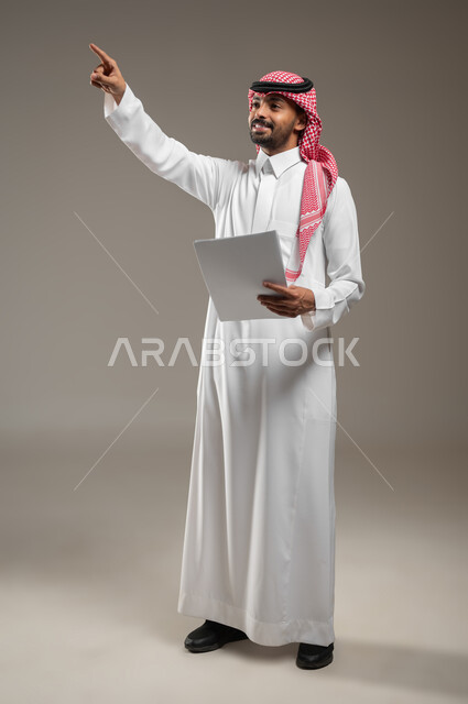 الإشارة إلى مكان ما والنظر إلى الاعلى, البشاشة والسرور وتحديد شيء معين, بورتريه بالطول الكامل لشاب عربي خليجي سعودي مبتسم يرتدي الثوب والشماغ السعودي يمسك بيده ورقه بيضاء ويشير بإصبع السبابة إلى شيء ما, ايماءات العثور على مكان ما، خلفيه رماديه، محتوى حساس