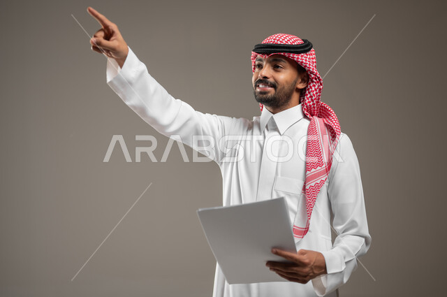 الإشارة إلى مكان ما والنظر إلى الاعلى, البشاشة والسرور وتحديد شيء معين, بورتريه مقرب لشاب عربي خليجي سعودي مبتسم يرتدي الثوب والشماغ السعودي يمسك بيده ورقه بيضاء ويشير بإصبع السبابة إلى شيء ما, ايماءات العثور على مكان ما، خلفيه رماديه، محتوى حساس