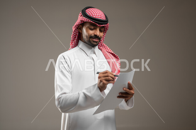 صورة مقربة جانبيه لرجل عربي خليجي سعودي يرتدي الزي السعودي يقوم بتدوين ملاحظاته على ورقه بيضاء، توقيع عقد ، شركة سعودية، عقود الموظفين، الملاحظات والسجلات، الرقابه الاداريه، موظف بدوام كامل، خلفيه رماديه، محتوى حساس