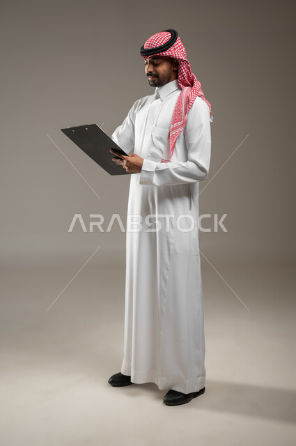 A Saudi Gulf Arab man in Saudi uniform, responsible for supervising work and monitoring the performance of employees, recording his observations and required tasks on a special file, taking notes and records, oversight and investigation, full-length side portrait, gray background, sensitive content