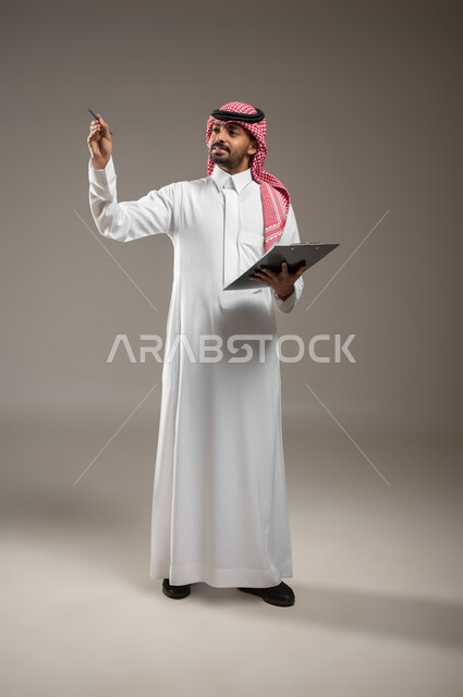 تدوين وتسجيل الملاحظات والأفكار، بورتريه بالطول الكامل لرجل عربي خليجي سعودي يرتدي الغترة والثوب السعودي يحمل بيده لوح تدوين الملاحظات، الكتابة والتخطيط على الأوراق والمستندات، يشير بقلمه الى مكان ما، تعابير وايماءات الشرح الانتباه والتركيز، خلفيه رماديه، محتوى حساس