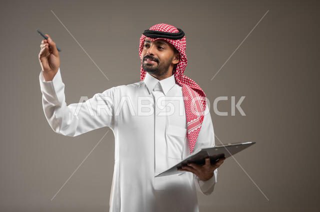 تدوين وتسجيل الملاحظات والأفكار، بورتريه مقرب لرجل عربي خليجي سعودي يرتدي الغترة والثوب السعودي يحمل بيده لوح تدوين الملاحظات، الكتابة والتخطيط على الأوراق والمستندات، يشير بقلمه الى مكان ما، تعابير وايماءات الشرح الانتباه والتركيز، خلفيه رماديهو محتوى حساس