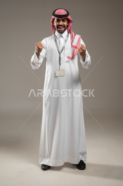 رفع اليد بإيماءات النصر والفوز, بورتريه لرجل عربي خليجي سعودي مبتسم يرتدي الزي السعودي ويضع بطاقه تعريفيه حول رقبته ينظر إلى الكاميرا بحماس, التفاعل والاندماج مع الألعاب الرياضية, الاحساس بالنشوه والنصر، صورة كامل الجسم، خلفية رمادية، محتوى حساس