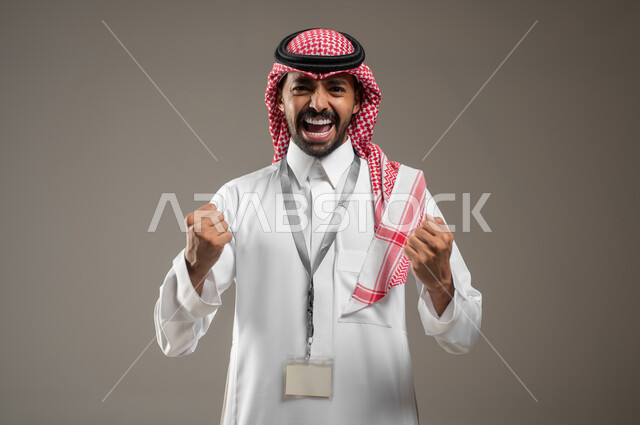 رفع اليد بإيماءات النصر والفوز, بورتريه مقرب لرجل عربي خليجي سعودي مبتسم يرتدي الزي السعودي ويضع بطاقه تعريفيه حول رقبته ينظر إلى الكاميرا بحماس, التفاعل والاندماج مع الألعاب الرياضية, الاحساس بالنشوه والنصر، خلفية رمادية، محتوى حساس