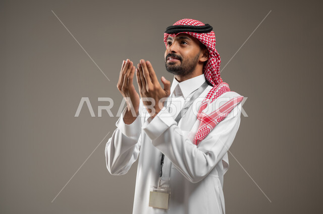 اشارات وجه ويدين تدل على الدعاء، التضرع الى الله, بورتريه جانبي مقرب لرجل عربي خليجي سعودي يرتدي الزي السعودي ويضع البطاقه التعريفيه حول رقبته، ينظر الى الاعلى، الرغبه في تحقيق الأمنيات, يرفع يديه للاعلى، يطلب العون والمغفره من الله، خلفيه رماديه، محتوى حساس