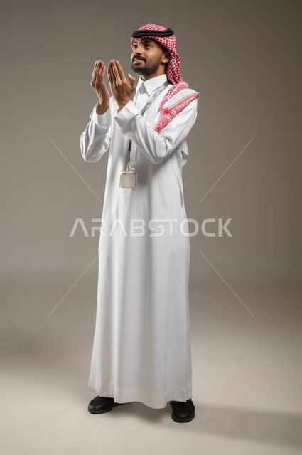 اشارات وجه ويدين تدل على الدعاء، التضرع الى الله, بورتريه جانبي بالطول الكامل لرجل عربي خليجي سعودي يرتدي الزي السعودي ويضع البطاقه التعريفيه حول رقبته، ينظر الى الاعلى، الرغبه في تحقيق الأمنيات, يرفع يديه للاعلى، يطلب العون والمغفره من الله، خلفيه رماديه، محتوى حساس