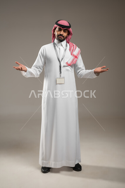 التساؤل والاستفهام والاستفسار عن شيء ما، ايماءات عدم المعرفة والدهشة والاستغراب، بورتريه  بالطول الكامل لرجل عربي خليجي سعودي يرتدي الشماغ والثوب التقليدي يرتدي البطاقه التعريفيه يرفع يديه للاعلى، ايماءات الشرح والتوضيح، خلفية رمادية، محتوى حساس