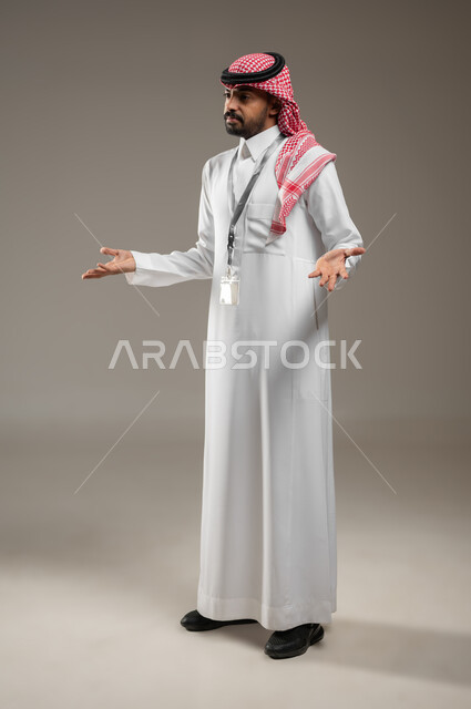 التساؤل والاستفهام والاستفسار عن شيء ما، ايماءات عدم المعرفة والدهشة والاستغراب، بورتريه جانبي بالطول الكامل لرجل عربي خليجي سعودي يرتدي الشماغ والثوب التقليدي يرتدي البطاقه التعريفيه يرفع يديه للاعلى، ايماءات الشرح والتوضيح، خلفية رمادية، محتوى حساس
