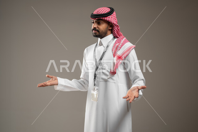Questioning, questioning, and inquiring about something, gestures of not knowing, astonishment, and astonishment, close side portrait of a Saudi Gulf Arab man wearing a shemagh and traditional dress, wearing an identification card, raising his hands up, gestures of explanation and clarification, gray background, sensitive content.