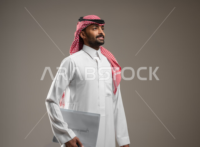 بورتريه جانبي مقرب لرجل عربي خليجي سعودي يرتدي الزي السعودي يحمل بيده موك اب ابيض، اوراق وملفات العمل، متابعة المراسلات والسجلات ،انجاز المهام المطلوبة، ينظر الى شئ ما بابتسامه، خلفيه رماديه، استخدام حساس، محتوى حساس