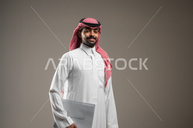 بورتريه جانبي مقرب لرجل عربي خليجي سعودي يرتدي الزي السعودي يحمل بيده موك اب ابيض، اوراق وملفات العمل، متابعة المراسلات والسجلات ،انجاز المهام المطلوبة، ينظر الى الكاميرا بابتسامه وثقه، خلفيه رماديه، استخدام حساس، محتوى حساس