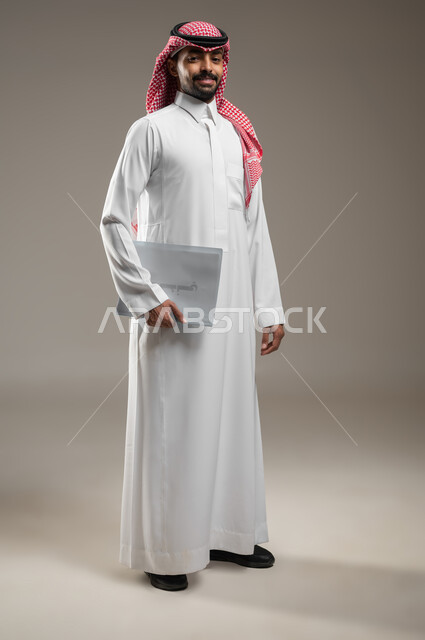 بورتريه جانبي بالطول الكامل لرجل عربي خليجي سعودي يرتدي الزي السعودي يحمل بيده موك اب ابيض، اوراق وملفات العمل، متابعة المراسلات والسجلات ،انجاز المهام المطلوبة، ينظر الى الكاميرا بابتسامه وثقه، خلفيه رماديه، استخدام حساس، محتوى حساس
