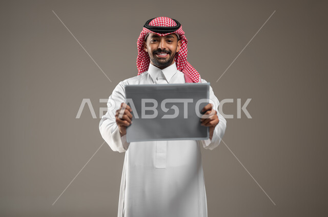 إعلانات وعروض تسويقية، استخدام لوحات توضيحية تعريفية، بورتريه مقرب لرجل عربي خليجي سعودي يرتدي الشماغ  والثوب السعودي يحمل بيده موك اب مربع رمادي ينظر إلى الكاميرا بايماءات السعاده، خلفية رمادية، محتوى حساس، استخدام حساس
