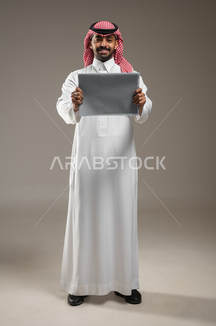 إعلانات وعروض تسويقية، استخدام لوحات توضيحية تعريفية، بورتريه لرجل عربي خليجي سعودي يرتدي الشماغ  والثوب السعودي يحمل بيده موك اب مربع رمادي ينظر إلى الكاميرا بايماءات السعاده، صورة الجسم بالكامل، خلفية رمادية، محتوى حساس، استخدام حساس