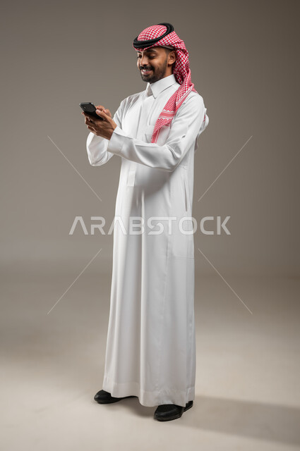 متابعة الاحداث العالمية باستخدام الجوال، بورتريه جانبي بالطول الكامل لشاب عربي سعودي خليجي يرتدي الزي السعودي يمسك بيده الهاتف المحمول، التواصل مع الأصدقاء عبر مواقع التواصل الاجتماعي، تقنيه الاتصالات، خلفيه رماديه ، استخدام حساس، محتوى حساس
