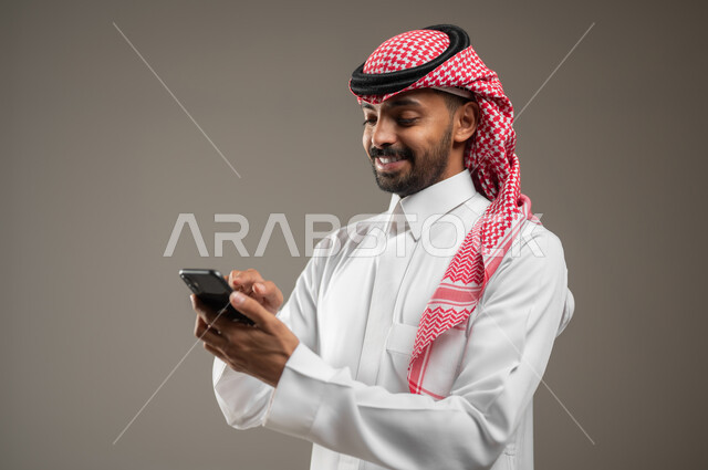 متابعة الاحداث العالمية باستخدام الجوال، بورتريه  مقرب جانبي لشاب عربي سعودي خليجي يرتدي الزي السعودي يمسك بيده الهاتف المحمول، التواصل مع الأصدقاء عبر مواقع التواصل الاجتماعي، تقنيه الاتصالات ونظم المعلومات، خلفيه رماديه ، استخدام حساس، محتوى حساس
