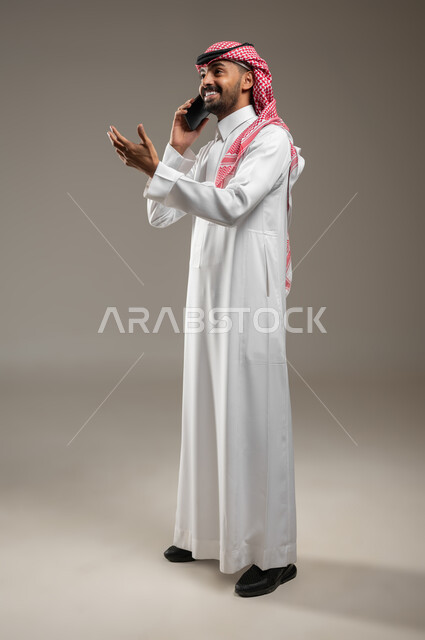 التواصل مع الأصدقاء والاقارب، الرد على الاتصالات والمكالمات، سماع أخبار سعيدة، بورتريه جانبي بالطول الكامل لرجل عربي سعودي خليجي يرتدي الشماغ والثوب السعودي يقوم بإجراء مكالمة هاتفية ويشير بيده نحو الاعلى بايماءات السعاده، خلفيه رماديه، محتوى حساس