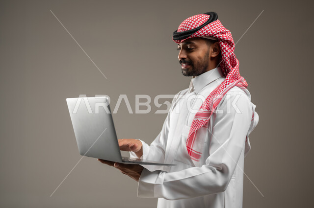 Browsing the Internet and social media programs, using a laptop to accomplish tasks, a close-up portrait of a Saudi Gulf Arab man wearing Saudi clothing holding a laptop, development of technology and technical devices, gray background, sensitive content