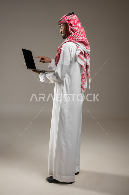 تصفح الانترنت وبرامج التواصل الاجتماعي, إستخدام الحاسوب المحمول لإنجاز المهام، بورتريه جانبي خلفي بالطول الكامل لرجل عربي سعودي خليجي يرتدي الزي السعودي يحمل جهاز اللابتوب(الكمبيوتر المحمول)، تطور التكنولوجيا والأجهزة التقنية، خلفية رمادية، محتوى حساس