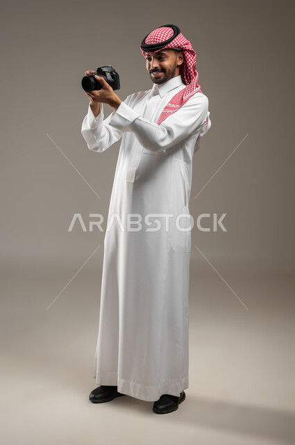 مصور فوتوغرافي يقوم بمراجعة لقطاته, بورتريه جانبي بالطول الكامل لشاب عربي خليجي سعودي مبتسم يرتدي الزي السعودي يحمل بيده كاميرا التصوير عالية الدقة، ينظر الى الصور الملتقطه بإيماءات الإعجاب والسعادة, معدات وتقنيات متطورة, خلفية رماديه، محتوى حساس