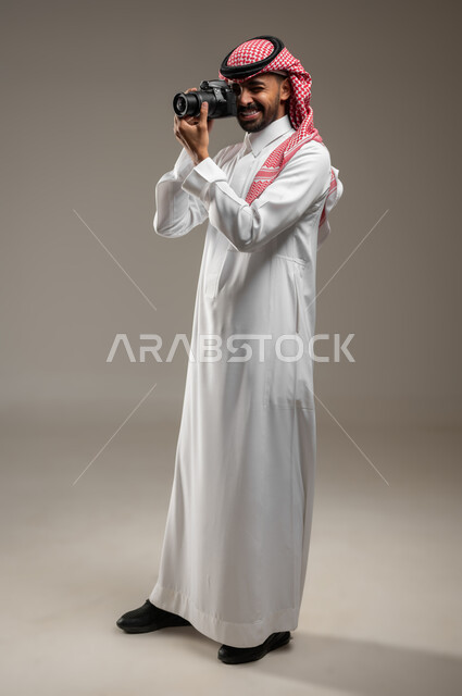 Using high-resolution digital cameras, a full-length side portrait of a professional Saudi Arabian Gulf photographer wearing the Saudi official uniform, taking souvenir photos with a photographic camera, practicing the hobby of photography, gray background, sensitive content