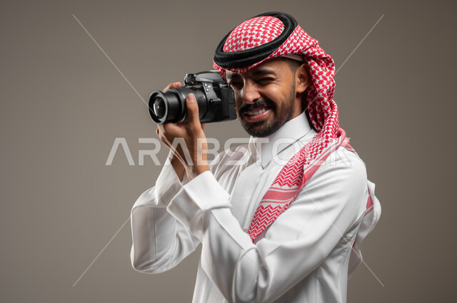استخدام كاميرات رقمية عالية الدقة, بورتريه مقرب من الجانب لمصور محترف عربي خليجي سعودي يرتدي الزي الرسمي السعودي يقوم بالتقاط صور تذكارية عبر كاميرا فوتوغرافية, ممارسة هواية التصوير, خلفية رماديه، محتوى حساس