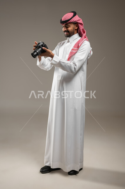 Enjoying practicing the hobby of photography, using a high-resolution photographic camera, a full-length side portrait of a young Saudi Arabian Gulf man wearing Saudi clothing, holding the camera in his hand and looking at it, documenting happy moments, gray background, sensitive content
