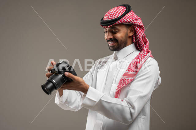 Enjoying practicing the hobby of photography, using a high-resolution photographic camera, a close-up portrait of a young Saudi Arabian Gulf man wearing Saudi clothing, holding the camera in his hand and looking at it, documenting happy moments, gray background, sensitive content