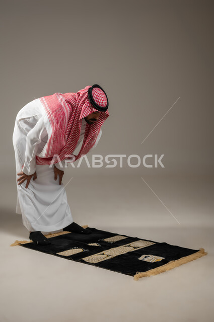 Performing the obligatory prayers on time, the second pillar of Islam, getting closer to God by performing the obligatory prayers and acts of worship, a full-length side portrait of a Saudi Gulf Arab man wearing the thobe, shemagh, and the Saudi headband, kneeling, gray background, sensitive use, sensitive content