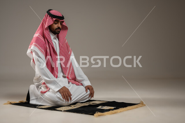 أهمية الالتزام بالصلاة على وقتها، بورتريه مقرب لرجل عربي سعودي خليجي يرتدي الثوب والشماغ والعقال السعودي، يجلس للتشهد يؤدي فريضه الصلاه على سجادته بخشوع، العبادة و التقرب الى الله، خلفية رماديه، محتوى حساس، استخدام حساس