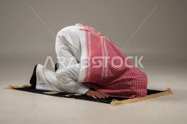 Supplication and humility during prostration, the second pillar of Islam, getting closer to God by performing the duties and acts of worship, a close-up portrait of a Saudi Gulf Arab man wearing the Saudi thobe and shemagh, prostrating, performing the obligatory prayer on time, gray background, sensitive use, sensitive content