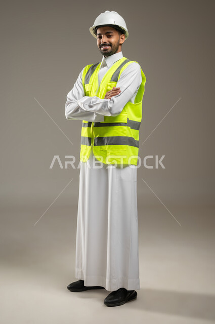 بورتريه جانبي بالطول الكامل لمهندس عربي خليجي سعودي يرتدي خوذة السلامة وسترة الحماية الخاصة بالعمل ، مهن ووظائف هندسية ، ادارة المشاريع الهندسية، مهندس معماري محترف، ينظر الى الكاميرا بابتسامه مكتوف اليدين، خلفيه رماديه، محتوى حساس، استخدام حساس