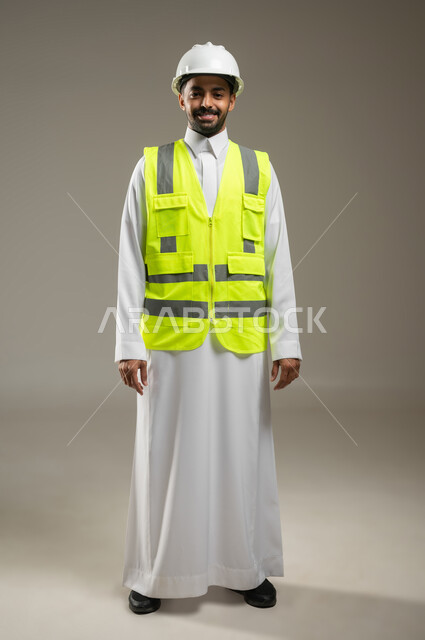Portrait of a Saudi Gulf Arab engineer wearing a safety helmet and work protection vest, engineering professions and jobs, engineering project management, professional architect, looking at the camera with a smile, full body photo, gray background, sensitive content, sensitive use
