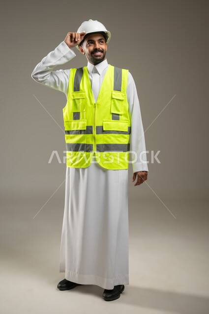 الفن في تقنية البناء والتصميم، متابعة سير المشاريع الإعمارية ، بورتريه لمهندس عربي خليجي سعودي يرتدي الخوذه و سترة الحماية،  يمسك بيده خوذه الحمايه بإيماءات السرور، ينظر الى شئ ما بابتسامه، صورة الجسم بالكامل، خلفية رمادية، محتوى حساس