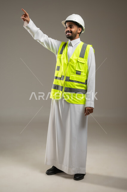 The concept of engineering and architectural construction, a full-length portrait of a Saudi Arabian Gulf engineer wearing the Saudi uniform, a protective jacket and helmet for engineering work, pointing with his index finger at something, gray background, sensitive content, sensitive use