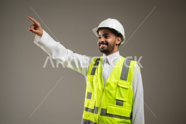 مفهوم الهندسة والانشاءات المعماريه،  بورتريه مقرب لمهندس عربي خليجي سعودي يرتدي الزي السعودي، سترة وخوذه الحمايه الخاصه بالعمل الهندسي، يشير باصبع السبابه الى شيء ما، خلفية رماديه, محتوى حساس، استخدام حساس