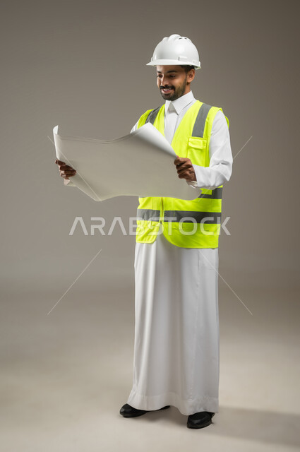 النظر إلى مخطط البناء، الحرص على بناء الأساسيات الجيده للمبنى ،التحقق من تطبيق الخطط و المشاريع الهندسيه المعمارية، مهندس عربي خليجي سعودي يرتدي الخوذة وسترة الحماية يعمل في القطاع الهندسي، يقوم بدراسه مخططات المشروع، بورتريه جانبي بالطول الكامل، خلفيه رماديه، محتوى حساس
