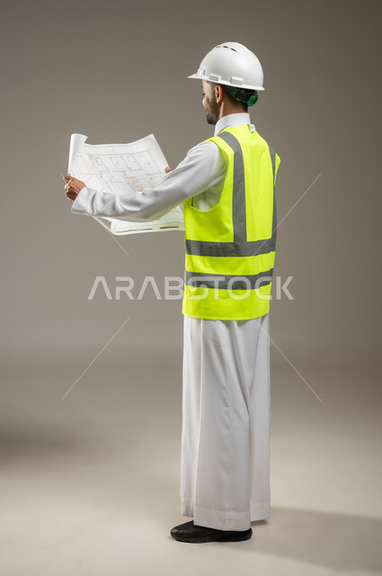 النظر إلى مخطط البناء، الحرص على بناء الأساسيات الجيده للمبنى ،التحقق من تطبيق الخطط و المشاريع الهندسيه المعمارية، مهندس عربي خليجي سعودي يرتدي الخوذة وسترة الحماية يعمل في القطاع الهندسي، يقوم بدراسه مخططات المشروع، بورتريه جانبي خلفي بالطول الكامل، خلفيه رماديه، محتوى حساس