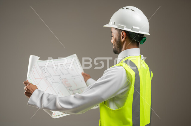 النظر إلى مخطط البناء، الحرص على بناء الأساسيات الجيده للمبنى ،التحقق من تطبيق الخطط و المشاريع الهندسيه المعمارية, مهندس عربي خليجي سعودي يرتدي الخوذة وسترة الحماية يعمل في القطاع الهندسي، يقوم بدراسه مخططات المشروع، بورتريه مقرب جانبي خلفي، خلفيه رماديه، محتوى حساس
