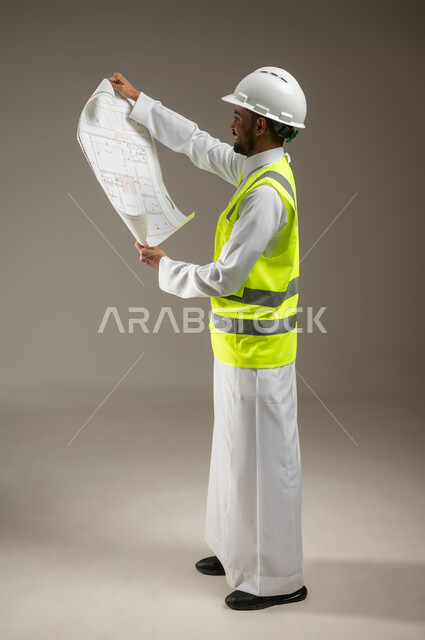 النظر إلى مخطط البناء، الحرص على بناء الأساسيات الجيده للمبنى ،التحقق من تطبيق الخطط و المشاريع الهندسيه المعمارية، مهندس عربي خليجي سعودي يرتدي الخوذة وسترة الحماية يعمل في القطاع الهندسي، يقوم بدراسه مخططات المشروع، بورتريه جانبي بالطول الكامل، خلفيه رماديه، محتوى حساس