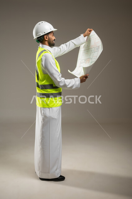 Supervision and control of engineering projects, construction with the help of the people of the nation, a full-length side portrait of a Saudi Gulf Arab engineer wearing the traditional dress with a helmet and protective jacket, holding the building plan and looking at it with pride, gray background, sensitive content, sensitive use