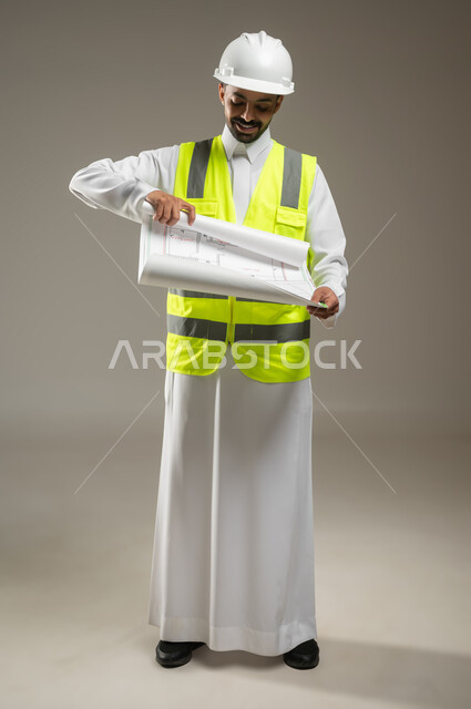الاشراف والرقابة على المشاريع الهندسية، الاعمار بسواعد ابناء الوطن، بورتريه  لمهندس عربي خليجي سعودي يرتدي الثوب التقليدي مع خوذة وسترة الوقاية يمسك مخطط البناء يقوم بفتحه وينظر إليه بفخر، صوره الجسم يالطول الكامل، خلفية رمادية، محتوى حساس، استخدام حساس